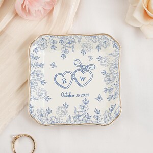 May include: A square, off-white ceramic ring dish with a gold rim and blue floral design. Two connected heart-shaped pendants with the letters "R" and "W" are in the center, with the date "October 25, 2025" below. A gold ring is in the foreground.