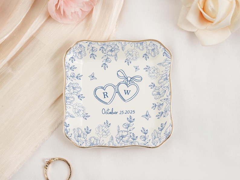 May include: A square, off-white ceramic ring dish with a gold rim and blue floral design. Two connected heart-shaped pendants with the letters "R" and "W" are in the center, with the date "October 25, 2025" below. A gold ring is in the foreground.