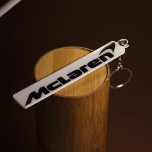 May include: A rectangular white acrylic keychain featuring the black McLaren logo and text printed on the front surface. The keychain includes a silver-toned metal jump ring and a circular split ring attached to the top right corner for keys.