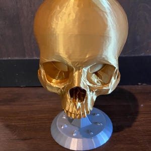 May include: A gold-colored skull-shaped decorative item. The skull has detailed facial features and sits atop a light gray, circular base. The item is likely 3D-printed and has a textured surface.