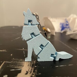 May include: A light blue, articulated dog keychain. The dog is segmented, allowing for movement, and features a textured surface. It is attached to a silver chain and clasp. The dog is sitting on a dark surface.