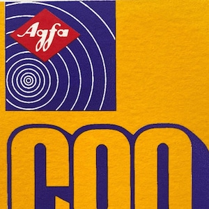 Könnte beinhalten: Ein Vintage-Etikett für eine Agfa C90 Kompaktkassette. Das Etikett ist gelb mit großem blauem und gelbem Text, der "C90" lautet. In der oberen linken Ecke befindet sich das Agfa-Logo in Rot und Weiß. Auf der Unterseite des Etiketts steht "Compact Cassette LOW-NOISE".