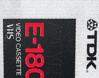 May include: A black and red E-180 VHS video cassette tape with the TDK logo. The tape has the words "HIGH OUTPUT HIGH RESOLUTION" printed at the top. The cassette is set against a white background.