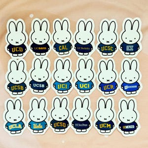 May include: A collection of white bunny stickers, each wearing a navy blue or blue sweater with a university logo. The stickers feature logos from UC Davis, UC Berkeley, UCLA, and other California universities.