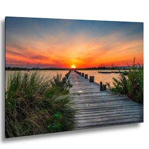 May include: A photograph of a sunset over a lake, featuring a wooden pier stretching into the water. The sun sets in a fiery sky of orange and red. Boats are visible in the distance. The pier is made of wooden planks.