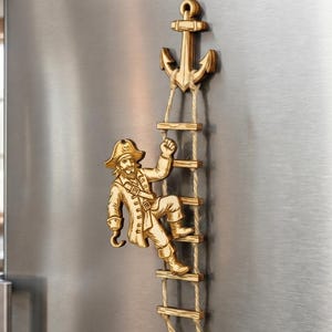 May include: Wooden pirate-themed refrigerator magnet. The magnet features a pirate climbing a rope ladder attached to an anchor. The pirate is wearing a hat and coat. The ladder is made of wood and rope.