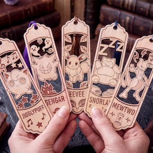 May include: Five light-colored wooden bookmarks featuring laser-cut designs of Jigglypuff, Gengar, Eevee, Snorlax, and Mewtwo. Each bookmark includes a colored tassel at the top and engraved character names with thematic icons like musical notes, ghosts, and Zs.