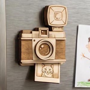 May include: Wooden camera-shaped magnet with a flash, lens, and a photo of a cat. The magnet is made of light-colored wood and is attached to a gray surface. A child's drawing is visible on the right.