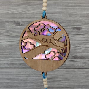 May include: A wooden, circular ornament featuring a laser-cut design of an airplane flying through clouds. The clouds and the plane's interior have a holographic, iridescent finish. The ornament is suspended by a blue string and wooden beads.