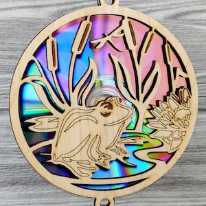 May include: Wooden circular ornament featuring a laser-cut design of a frog, cattails, and a dragonfly. The background is a colorful, iridescent CD. The ornament has two small holes at the top for hanging.