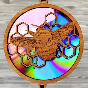May include: A circular wooden ornament featuring a laser-cut bee and honeycomb design. The ornament has a natural wood finish and a colourful, iridescent background. It includes a small hole for hanging, suitable for decorative purposes.