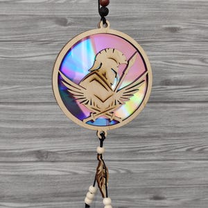 Puede incluir: Adorno circular de madera con un diseño de casco espartano y alas. El adorno tiene un fondo iridiscente. Cuelga de un cordón negro con cuentas y una pluma decorativa.