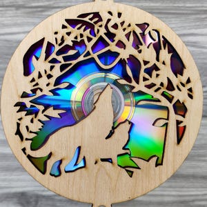 Puede incluir: Adorno redondo de madera con un diseño cortado con láser que presenta la silueta de un lobo aullando sobre un CD. El adorno tiene un acabado de madera natural y un patrón de árbol en los bordes. El CD refleja tonos azules, verdes y morados.
