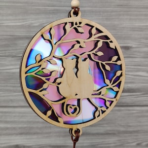 Puede incluir: Un móvil de viento de madera con silueta de gato. El diseño presenta dos gatos en un árbol, con un corazón debajo. El fondo es un material iridiscente y colorido. El móvil es redondo y cuelga de un cordón de cuero.