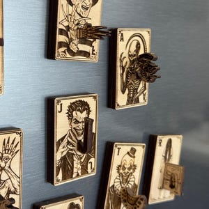 May include: Wooden playing card-shaped magnets featuring horror movie characters. Each magnet has a unique design, including Freddy Krueger, the Joker, and a skeletal figure. The magnets are brown with dark brown accents.
