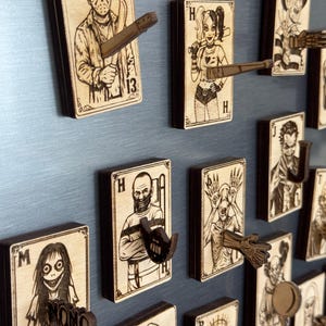 May include: Wooden playing card-shaped wall hooks featuring horror movie characters. Each hook has a unique design, including Jason Voorhees, Harley Quinn, and Freddy Krueger. The hooks are made of light-colored wood with dark brown edges.