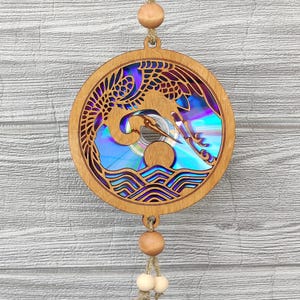 May include: Wooden circular wall hanging featuring a laser-cut design of a crane in flight over waves. The center of the circle has a holographic effect. The piece is suspended by a natural fiber cord with wooden beads.