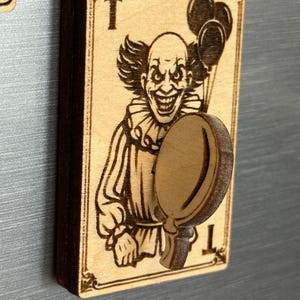 May include: A wooden magnet featuring a detailed engraving of a sinister clown holding balloons and a mirror. The design is on a playing card with the letter "T" at the top and bottom. The magnet is made of light brown wood.