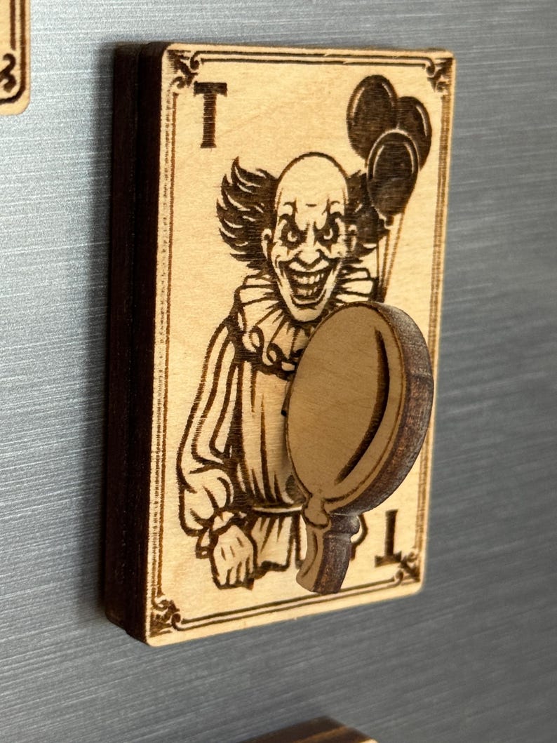 May include: A wooden magnet featuring a detailed engraving of a sinister clown holding balloons and a mirror. The design is on a playing card with the letter "T" at the top and bottom. The magnet is made of light brown wood.