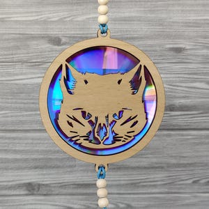 May include: A wooden cat-shaped ornament with a laser-cut design. The cat's face is in the centre of a circular frame. The centre of the ornament has a colourful, iridescent material. The ornament is suspended by a string of wooden beads and blue cord.