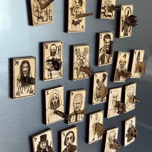 Può includere: Una collezione di magneti a forma di carte da gioco in legno con personaggi di film horror. Ogni carta presenta un'illustrazione unica e un piccolo accessorio in legno. Le carte sono disposte su una superficie azzurra.