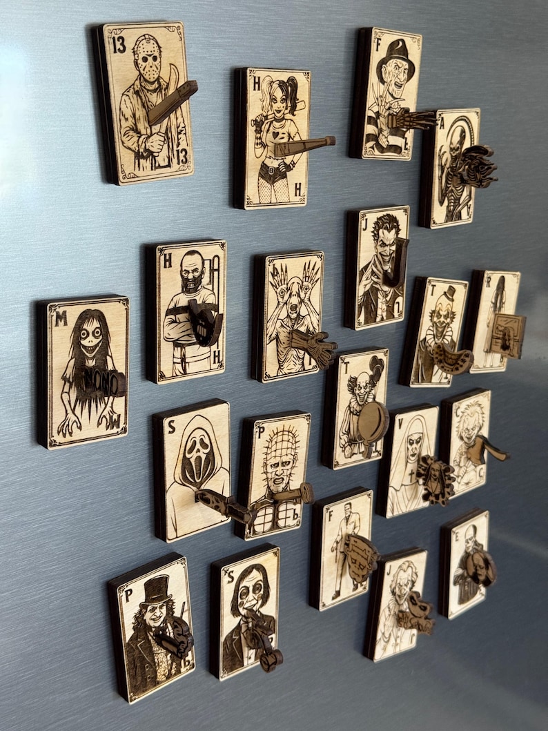 May include: A collection of wooden playing card magnets featuring horror movie characters. Each card has a unique illustration and a small, wooden prop extending from the card. The cards are arranged on a light blue surface.