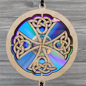 May include: A wooden Celtic cross ornament with a CD center. The cross design is intricately carved, featuring interwoven knot patterns. The CD reflects a spectrum of colors, adding a vibrant contrast to the natural wood. The ornament has a black string for hanging.