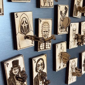 May include: Wooden playing card magnets featuring horror movie characters. Each card has a unique engraved design and a 3D element. The cards are displayed on a blue surface, showcasing the detailed craftsmanship and thematic collection.