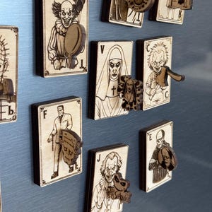 May include: Wooden playing card magnets featuring horror movie characters. Each magnet has a detailed illustration of a character, with a matching wooden accent piece. The magnets are displayed on a metallic surface.