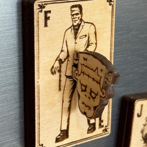 May include: Wooden magnet featuring a laser-etched image of Frankenstein's monster on a playing card. The card has the letter "F" at the top and a decorative border. A separate wooden head of the monster is attached to the card.