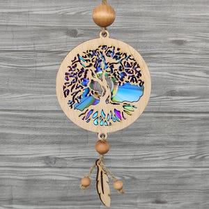 Op de afbeelding: Houten wanddecoratie met een levensboom met een lasergesneden ontwerp. De takken en bladeren van de boom zijn ingewikkeld gedetailleerd, met een kleurrijke, iriserende achtergrond. De hanger bevat houten kralen en een veerketting, opgehangen aan touw.