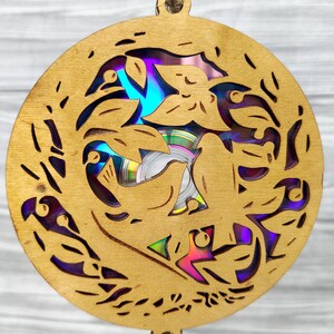 May include: A round, wooden ornament with a laser-cut design featuring birds and leaves. The ornament has a natural wood finish and a hole at the top for hanging. The center reveals a colorful, iridescent CD. The overall theme is nature and art.