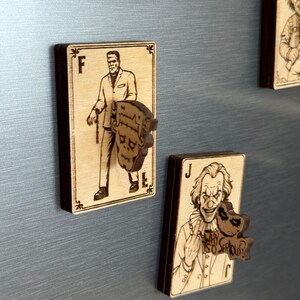 May include: Wooden playing card magnets featuring classic horror movie characters. The "F" card shows Frankenstein, while the "J" card features the Joker with the text "WHY SO SERIOUS?". The magnets are light brown with dark brown edges.