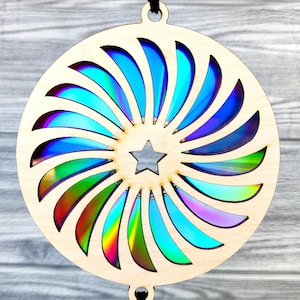 May include: A wooden, circular suncatcher with a star cutout in the center. The design features a spiral of iridescent, rainbow-colored segments. The suncatcher hangs from a black cord, creating a decorative and colorful accent.