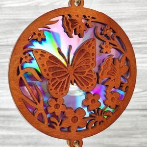 May include: A wooden butterfly ornament with a laser-cut design. The butterfly is the focal point, surrounded by flowers and foliage. The ornament is circular and has a CD in the center, with a rainbow iridescent effect. The wood is a warm brown color.