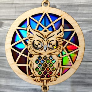 May include: Wooden owl ornament with a laser-cut design. The owl is centered within a circular frame featuring a geometric pattern with iridescent, rainbow-colored accents. A natural twine loop is attached for hanging.