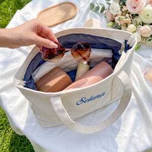 K&ouml;nnte beinhalten: Eine cremefarbene Canvas-Tragetasche mit dem in blauem Garn aufgestickten Namen Selena auf der Vorderseite. Das Innere ist mit dunkelblauem Stoff ausgekleidet und enth&auml;lt ein beiges rechteckiges Etui, eine rosa Flasche und ein kleines Fl&auml;schchen.