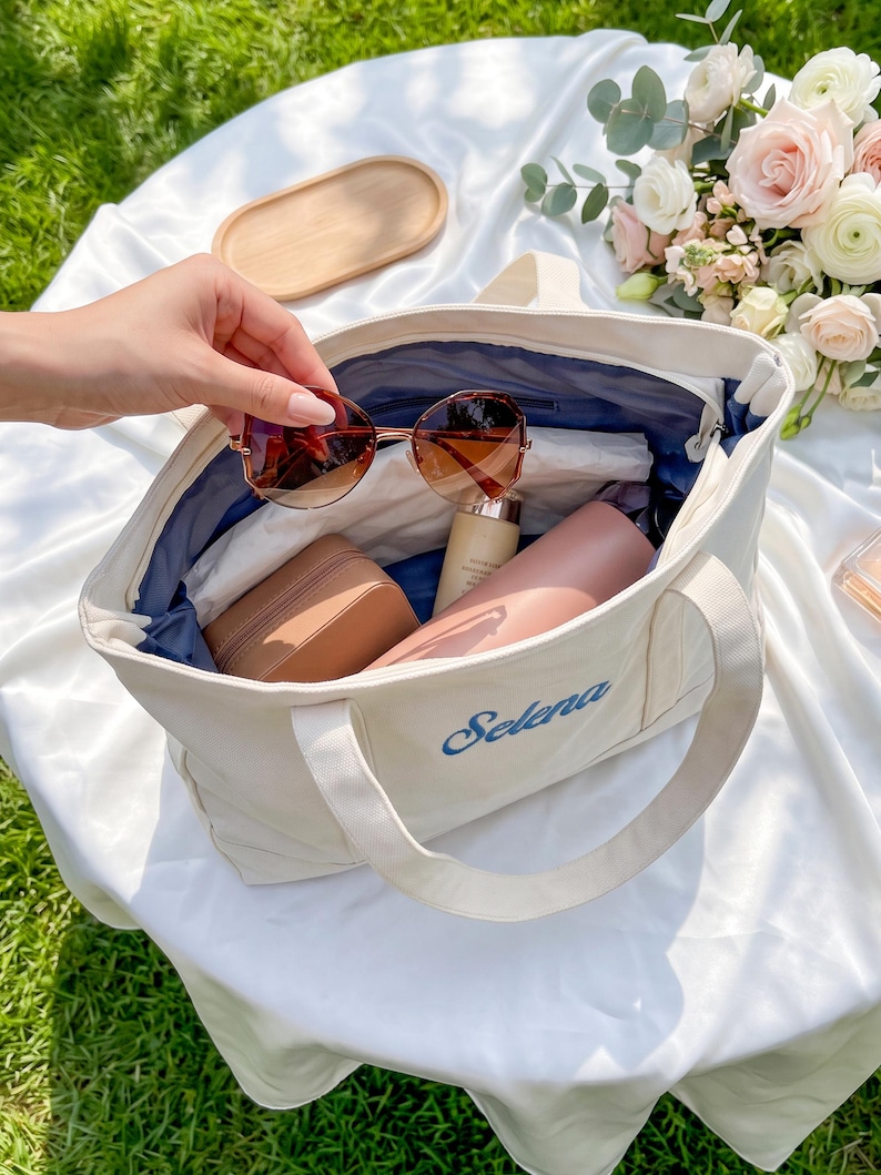 K&ouml;nnte beinhalten: Eine cremefarbene Canvas-Tragetasche mit dem in blauem Garn aufgestickten Namen Selena auf der Vorderseite. Das Innere ist mit dunkelblauem Stoff ausgekleidet und enth&auml;lt ein beiges rechteckiges Etui, eine rosa Flasche und ein kleines Fl&auml;schchen.