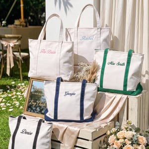 K&ouml;nnte beinhalten: F&uuml;nf cremefarbene Canvas-Tragetaschen mit Doppelgriffen und gestickten Namen oder Titeln. Eine Tasche zeigt den rosa Schriftzug 'The Bride', eine hellblaue 'Rachel', eine gr&uuml;ne 'Miss Smith', eine marineblaue 'Georgia' und eine schwarze 'Denise' mit passendem Besatz.