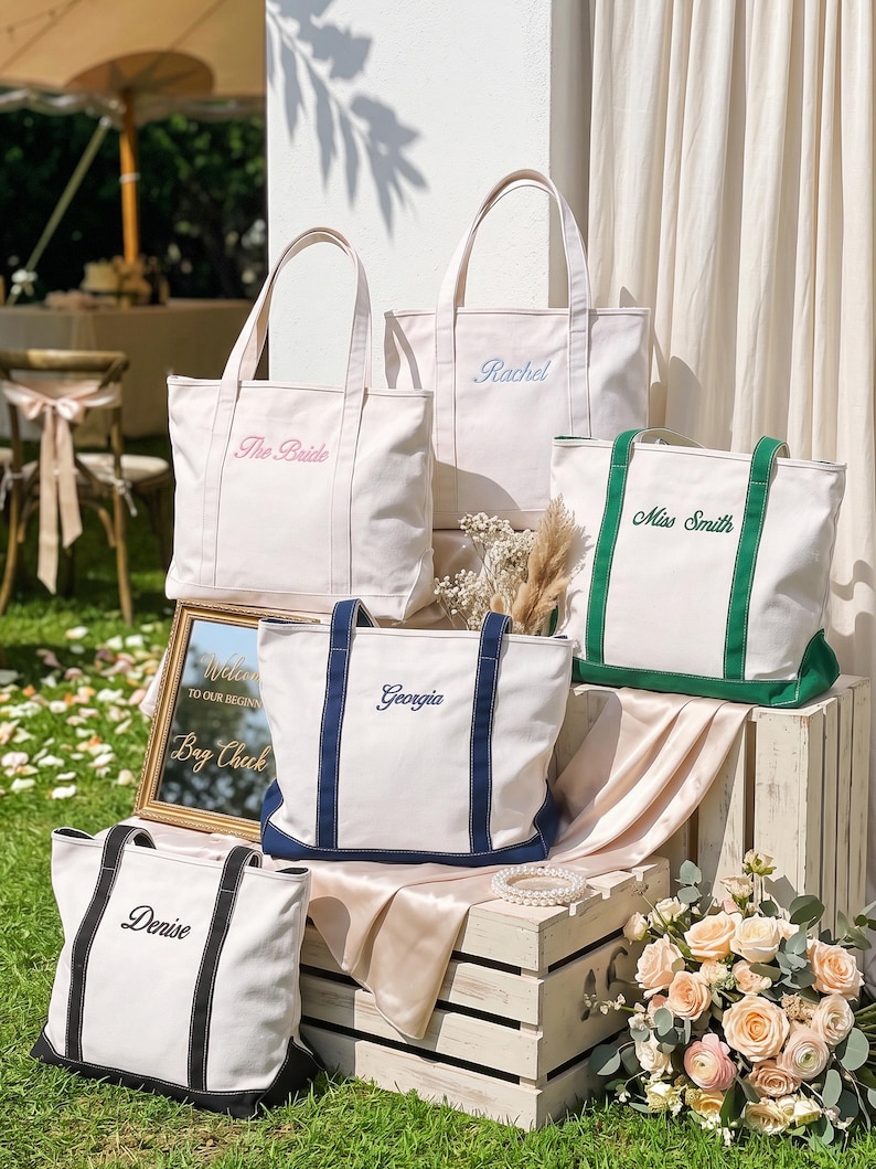 K&ouml;nnte beinhalten: F&uuml;nf cremefarbene Canvas-Tragetaschen mit Doppelgriffen und gestickten Namen oder Titeln. Eine Tasche zeigt den rosa Schriftzug 'The Bride', eine hellblaue 'Rachel', eine gr&uuml;ne 'Miss Smith', eine marineblaue 'Georgia' und eine schwarze 'Denise' mit passendem Besatz.