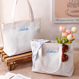 K&ouml;nnte beinhalten: Zwei cremewei&szlig;e Canvas-Tragetaschen mit zwei Henkeln. Eine Tasche ist mit dem Namen Natalie in blauer Schreibschrift bestickt, die andere mit dem Namen Selena in blauer Schreibschrift, einem blauen Blumenhalstuch und einer braunen Sonnenbrille.