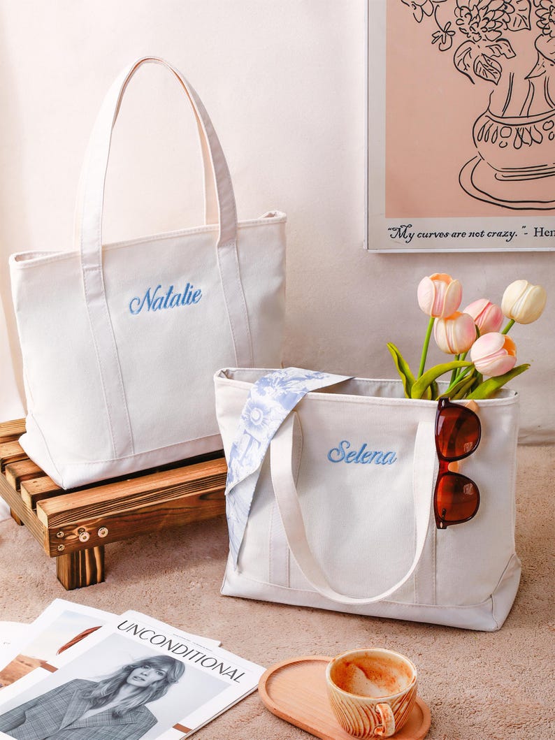K&ouml;nnte beinhalten: Zwei cremewei&szlig;e Canvas-Tragetaschen mit zwei Henkeln. Eine Tasche ist mit dem Namen Natalie in blauer Schreibschrift bestickt, die andere mit dem Namen Selena in blauer Schreibschrift, einem blauen Blumenhalstuch und einer braunen Sonnenbrille.