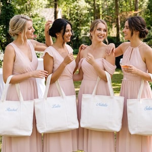 K&ouml;nnte beinhalten: Vier wei&szlig;e Tragetaschen aus Segeltuch, jede mit einem in hellblauer Schreibschrift gestickten Namen versehen. Die auf den Taschen angezeigten Namen sind Rachel, Laila, Natalie und Selena. Jede Tasche hat zwei stabile Griffe und einen rechteckigen Boden.