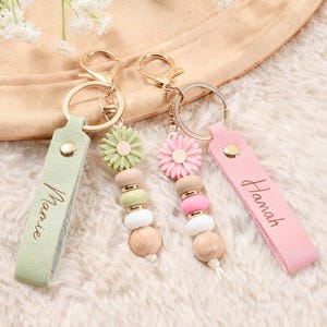 Op de afbeelding: Twee sleutelhangers met goudkleurige hardware. Eén sleutelhanger heeft een lichtgroene band met de naam "Mamie" en een bloemvormige bedel. De andere heeft een roze band met de naam "Hannah" en een bloemvormige bedel. Beide hebben houten kralen.