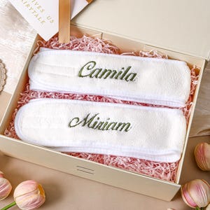Op de afbeelding: Een beige geschenkdoos met twee witte spa-hoofdbanden geborduurd met de namen "Camila" en "Miriam" in groene draad. De hoofdbanden liggen op roze papierversnippering. Een kaart met de tekst "Gives Us Fairytale" is ook zichtbaar.