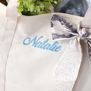 K&ouml;nnte beinhalten: Eine cremewei&szlig;e Canvas-Tragetasche mit dem in hellblauer Schreibschrift gestickten Namen Natalie. Ein gemustertes Stoffband mit schwarz-wei&szlig;em Aufdruck ist um den Griff gebunden, und gr&uuml;nes Blattwerk befindet sich im Inneren der Tasche.