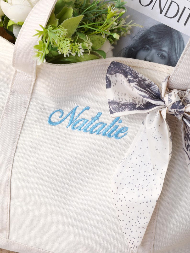 K&ouml;nnte beinhalten: Eine cremewei&szlig;e Canvas-Tragetasche mit dem in hellblauer Schreibschrift gestickten Namen Natalie. Ein gemustertes Stoffband mit schwarz-wei&szlig;em Aufdruck ist um den Griff gebunden, und gr&uuml;nes Blattwerk befindet sich im Inneren der Tasche.