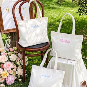 K&ouml;nnte beinhalten: Drei cremewei&szlig;e Tragetaschen aus Segeltuch mit jeweils zwei Henkeln. Eine Tasche zeigt den blau gestickten Namen Natalie, eine weitere den rosa gestickten Text The Bride und die dritte den blau gestickten Namen Selena auf der Vorderseite.