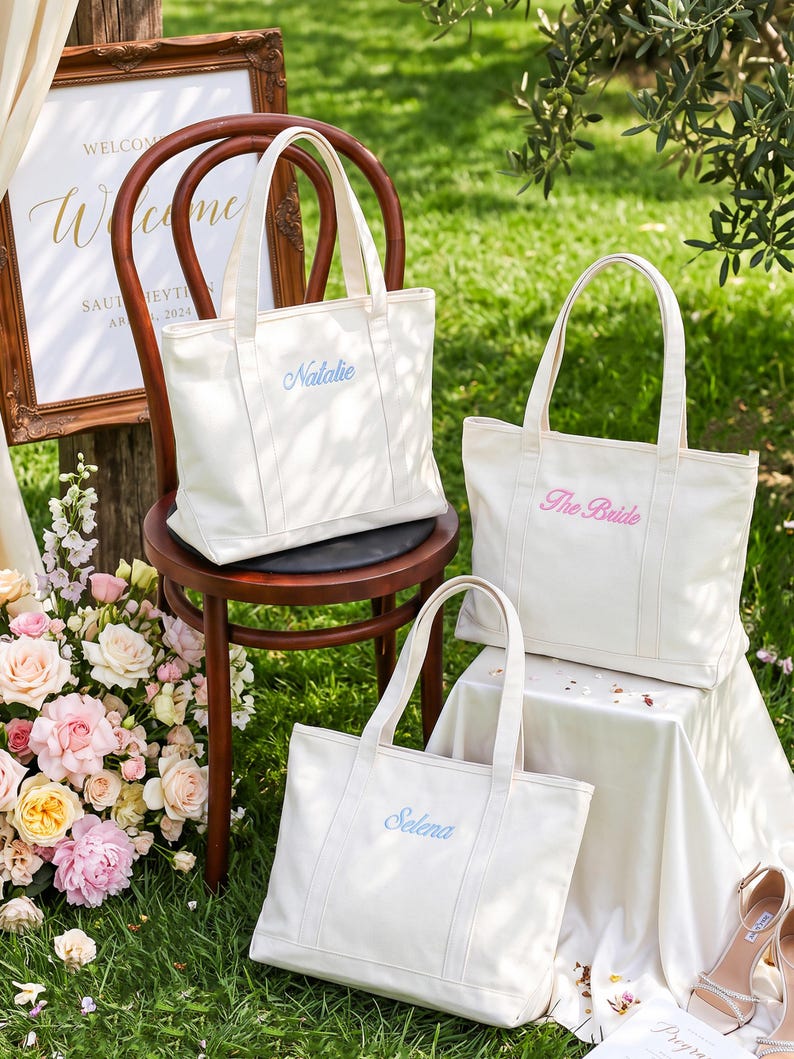 K&ouml;nnte beinhalten: Drei cremewei&szlig;e Tragetaschen aus Segeltuch mit jeweils zwei Henkeln. Eine Tasche zeigt den blau gestickten Namen Natalie, eine weitere den rosa gestickten Text The Bride und die dritte den blau gestickten Namen Selena auf der Vorderseite.