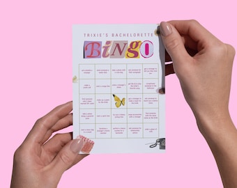 Könnte beinhalten: Eine Bingo-Karte für einen Junggesellinnenabschied mit dem Titel "Trixie's Bachelorette Bingo" mit farbenfrohem Text. Die Karte zeigt eine gelbe Schmetterlingsgrafik und verschiedene Aufforderungen. Die Karte wird vor einem rosa Hintergrund gehalten.